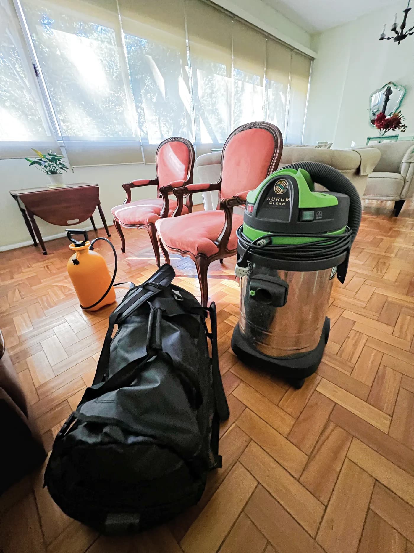 Sala com piso de parquet e duas poltronas rosa, com o aspirador da Aurum Clean, o pulverizador e a mochila da equipe no chão