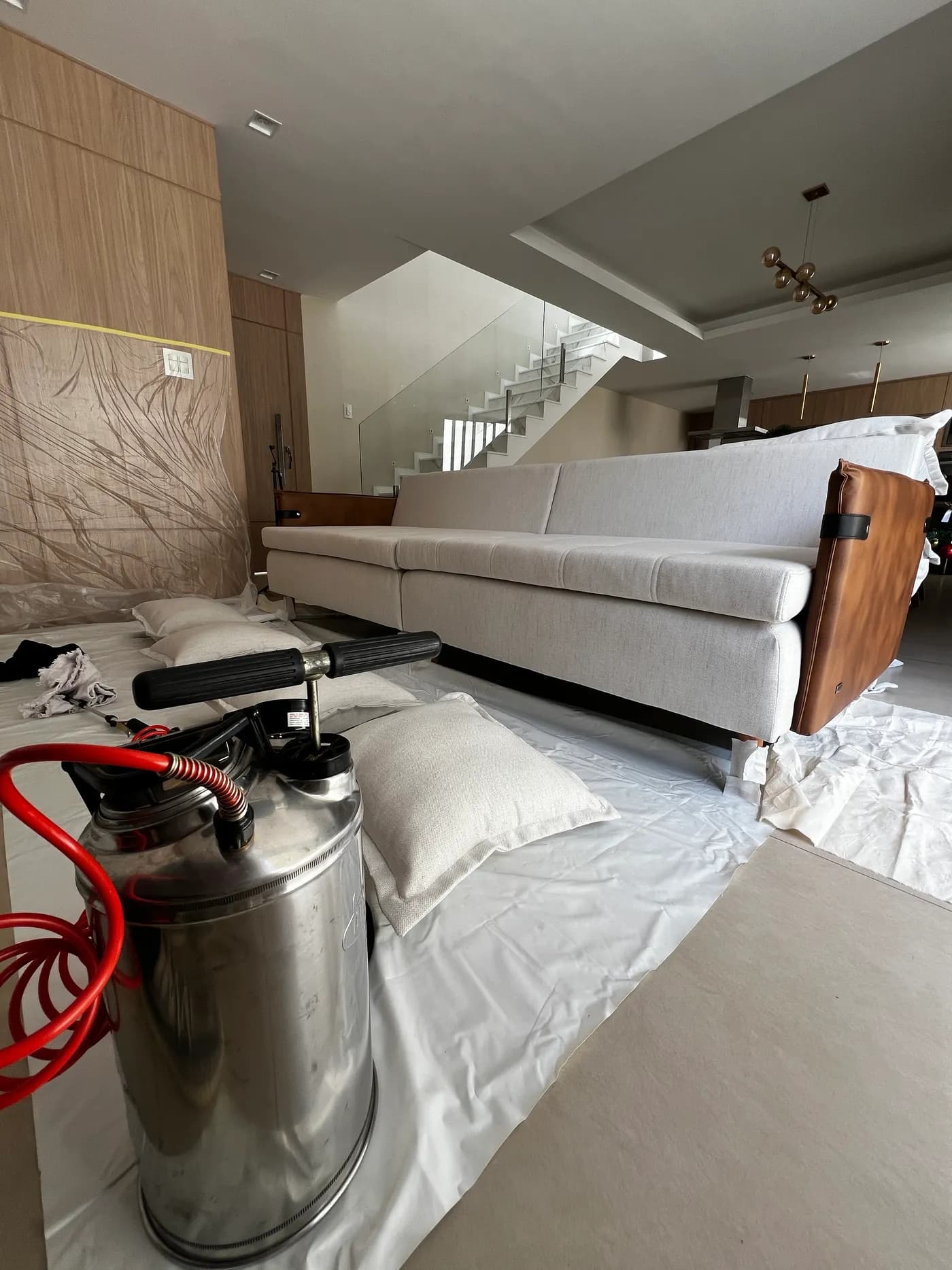 Sofá claro com lateral em couro caramelo, piso protegido com plástico e o pulverizador em primeiro plano